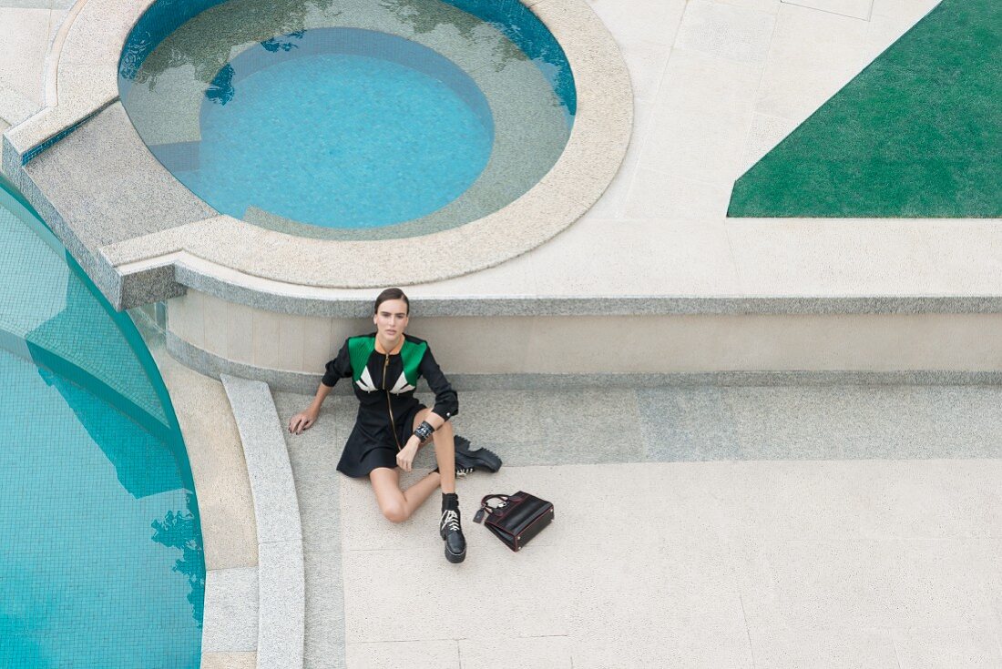 Brünette Frau in schwarzem Kleid mit Reißverschluss, Plateau-Stiefel und Henkeltasche am Pool