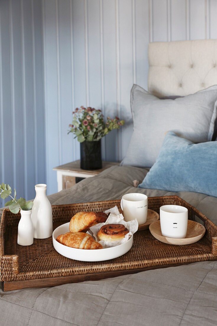 Pastries, cups and vase on wicker tray on grey bedspread