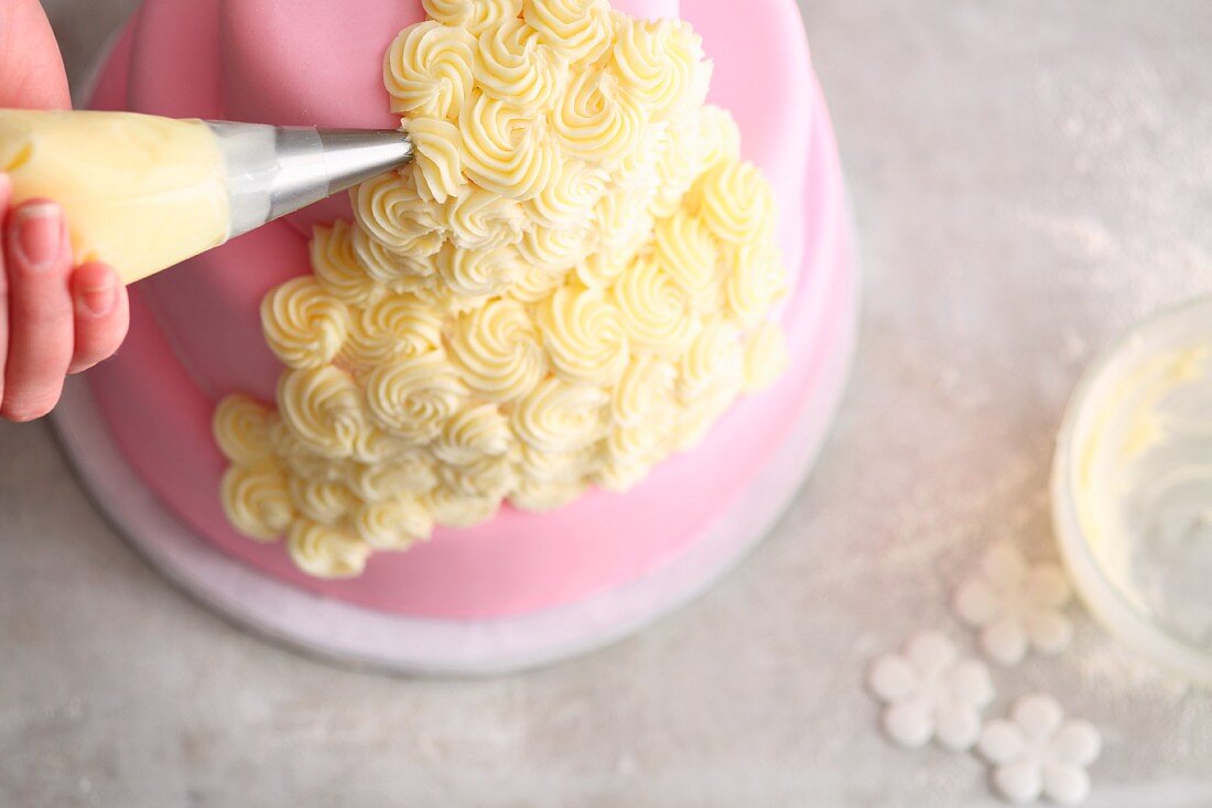 Three-tier pink fondant icing cake being decorated with buttercream