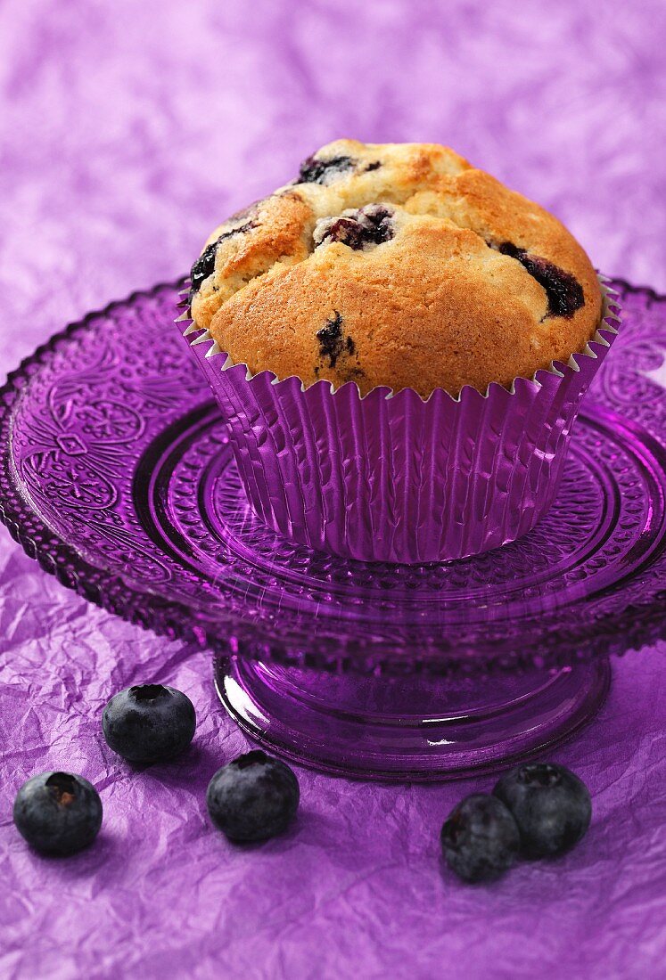 A blueberry muffin on a small purple glass cake stand sitting on a purple crushed paper background