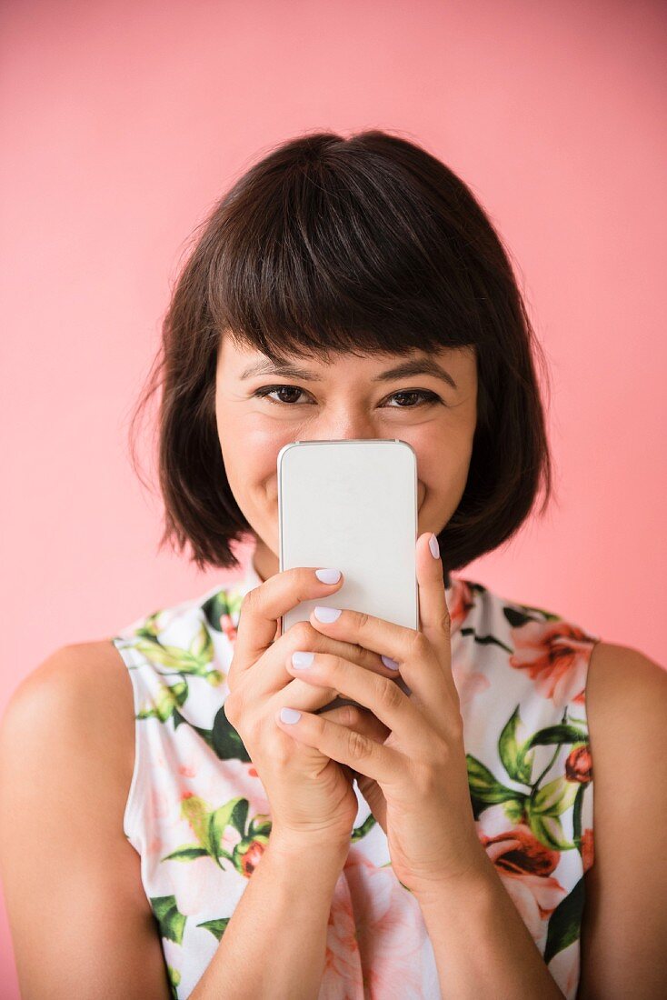 A young brunette woman wearing a floral dress and hiding her face behind a smartphone