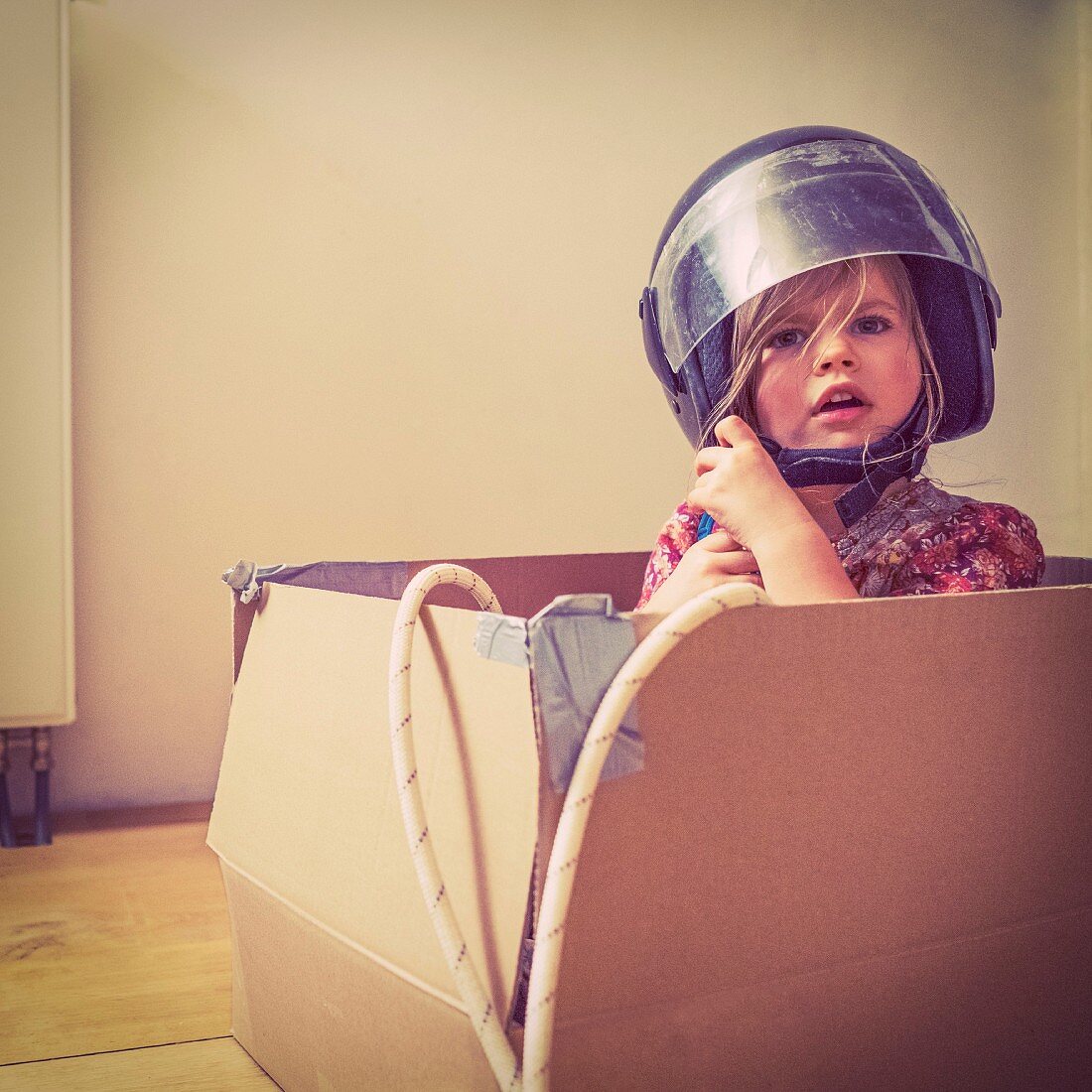 A little girl wearing a motorbike helmet sitting in a cardboard box