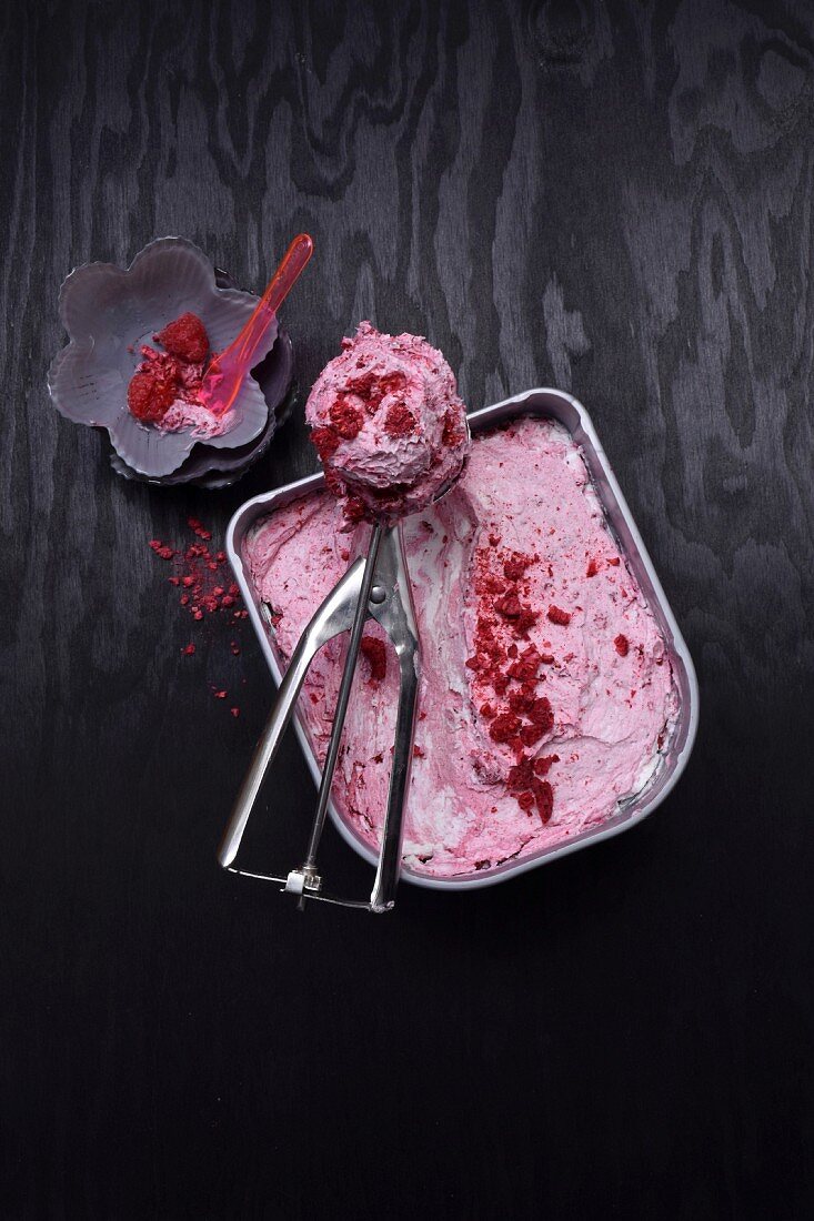 Raspberry and yoghurt ice cream in an ice cream container, an ice cream scoop, and a scoop of ice cream