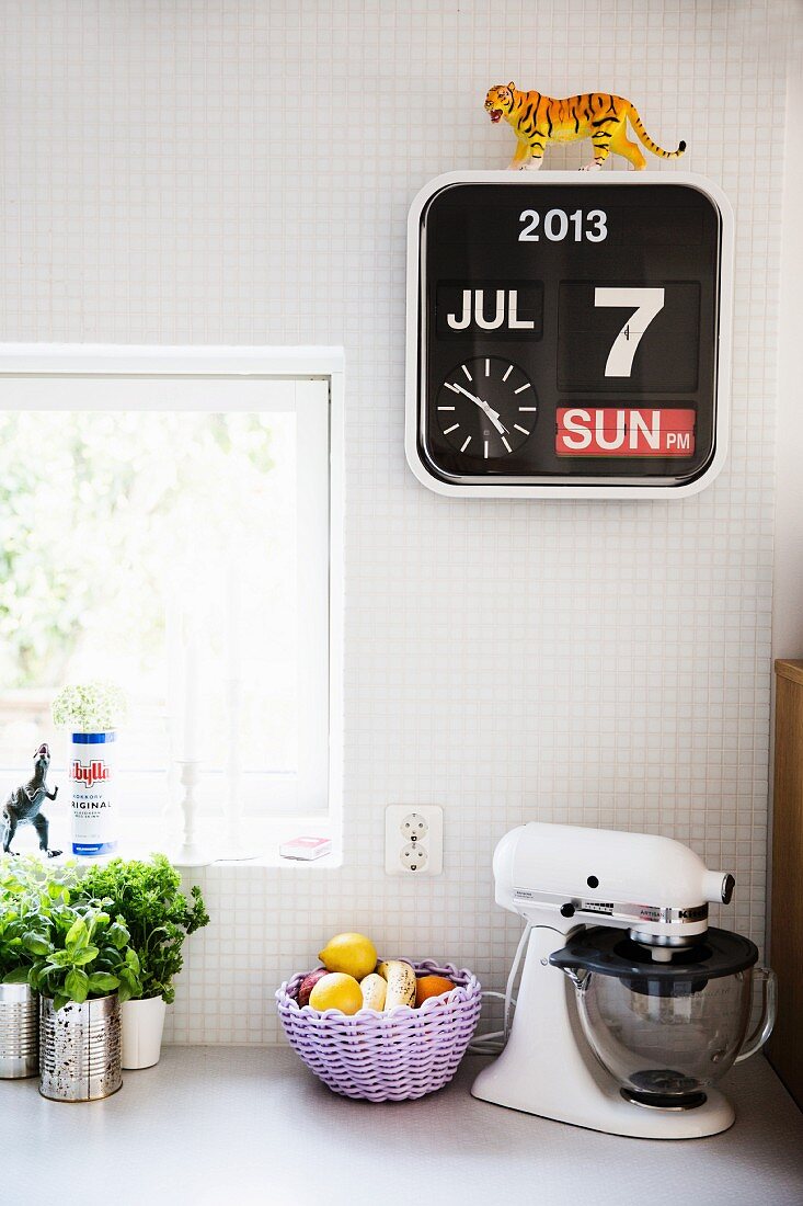 Herbs, fruit basket and mixer on kitchen counter below calendar clock on white-tiled wall