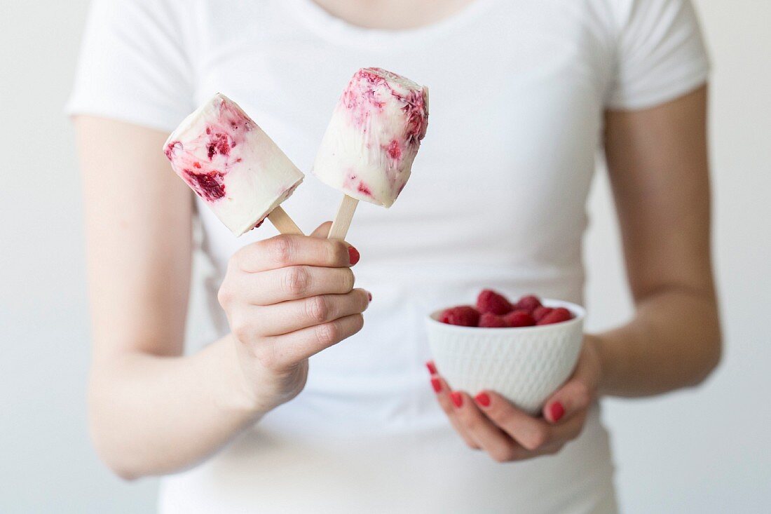A woman holding raspberry ice cream sticks and a bowl of fresh raspberries
