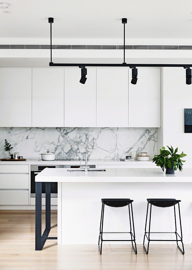 Black bar stools in front of a white kitchen island under a light rail with black spotlights in an open designer kitchen with marble splash protection
