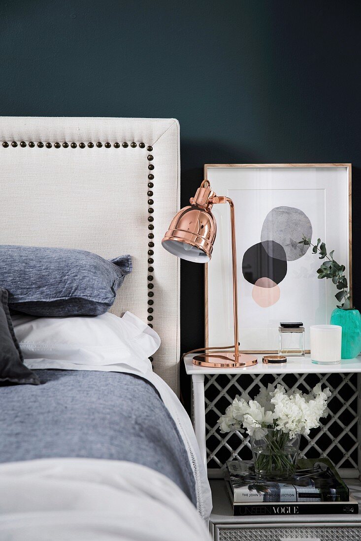 Weisses Bett mit Kopfteil neben Nachttisch mit kupferfarbener Tischleuchte vor schwarzer Wand