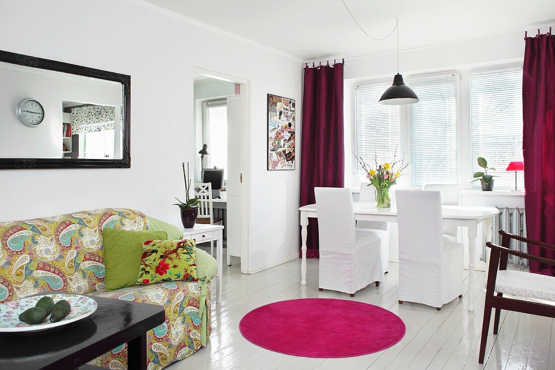 White, loose-covered chairs and dining table on white wooden floor, hot pink round rug and patterned sofa in open-plan living area