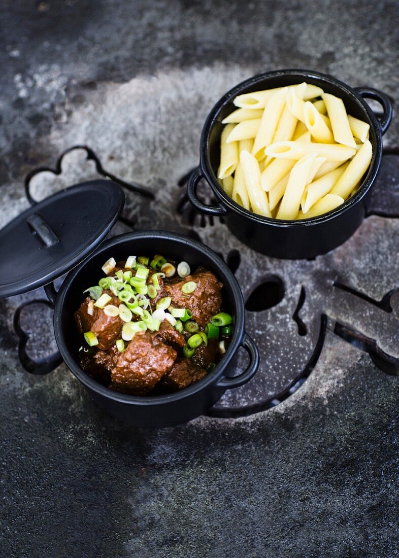 Goulash with spring onions and penne pasta in black pots