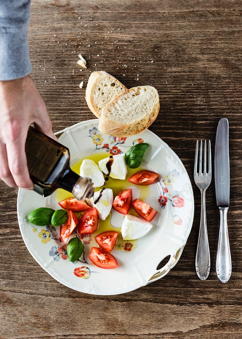 Balsamico auf Mozzarella, Tomatenstücke, Basilikum und Baguette auf altem Porzellanteller mit Blumendekor gießen