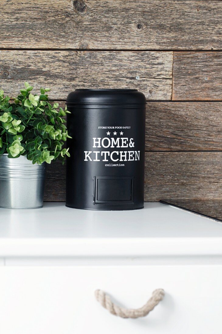 Schwarze Metalldose und Grünpflanze auf Küchenunterschrank vor rustikaler Holzverkleidung