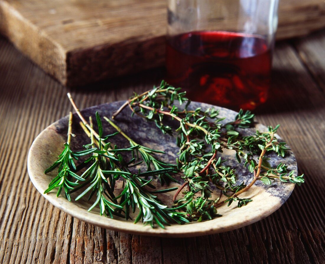 Sprigs of rosemary and thyme in a vintage bowl with a bottle of red wine in the background