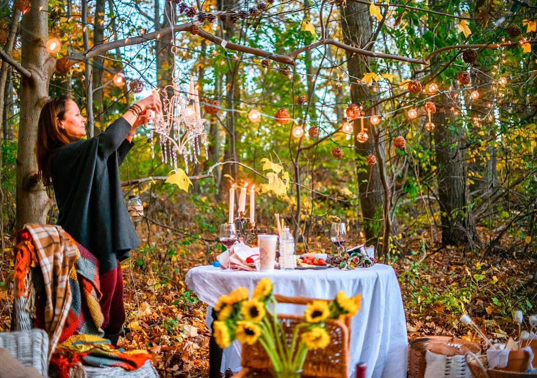 Frau dekoriert herbstlich gedeckten Tisch im Wald mit Lichterkette und Kristalleuchter