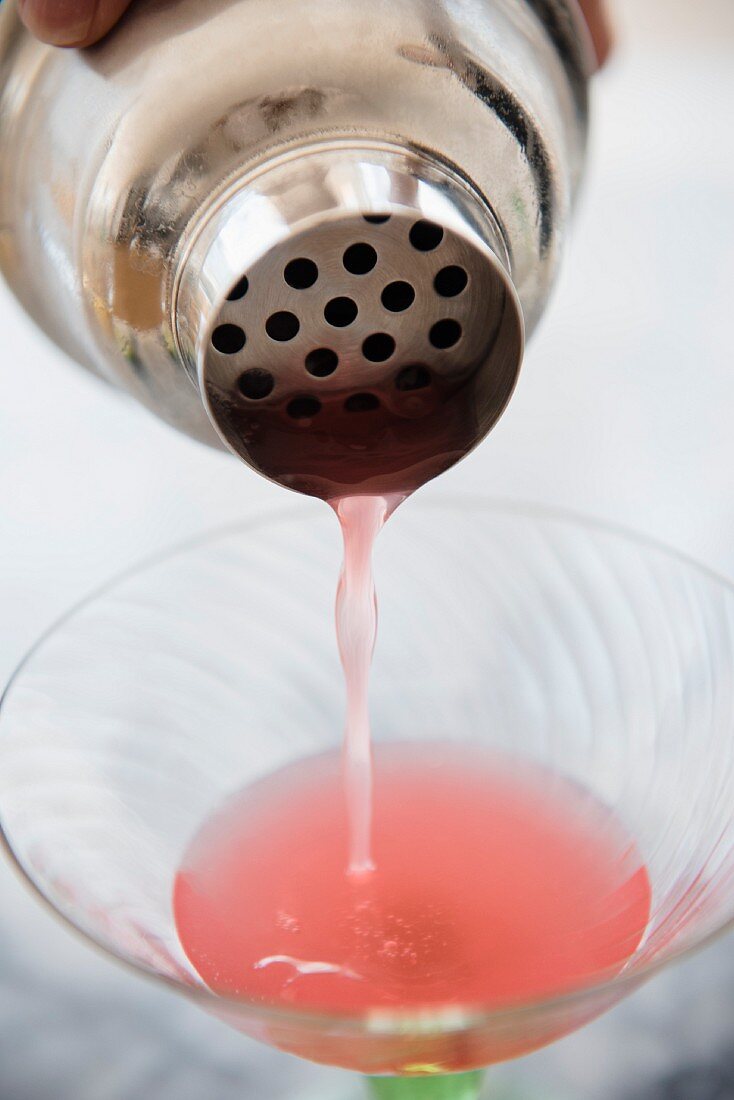 A pink cocktail being poured from a cocktail shaker into a martini glass