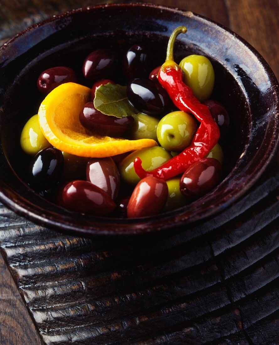 Green olives, black olives, a red chilli, an orange slice and a bay leaf in vintage bowl