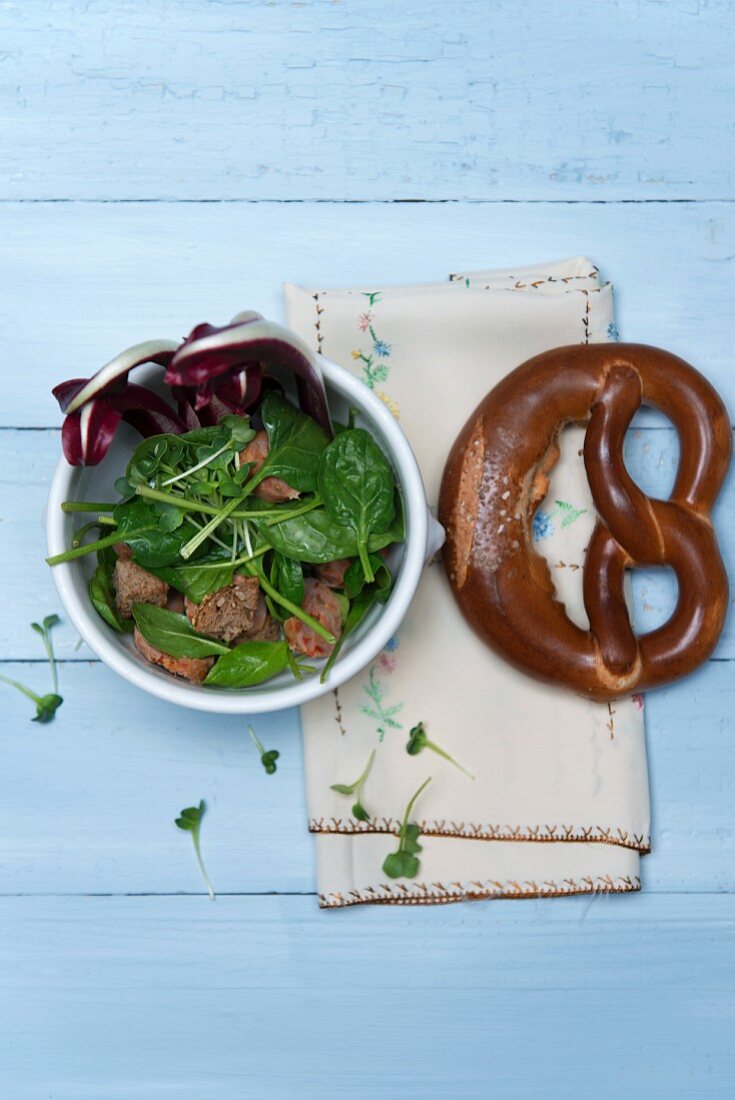 Spinach salad with chorizo, crostini, cress and Trevisio in a porcelain bowl next to a pretzel