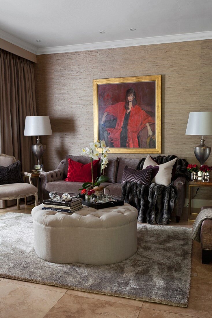 Round, beige ottoman on rug in front of sofa below gilt-framed portrait of woman on wallpapered wall