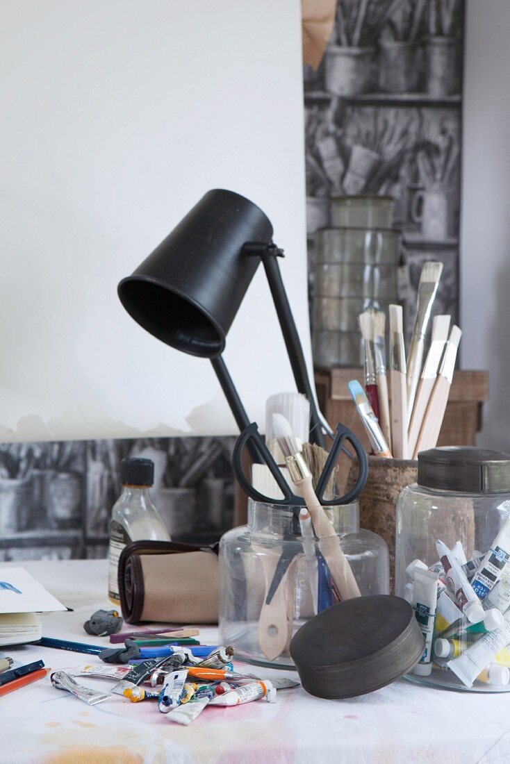 Artist's utensils in various storage jars, tubes of paint and black table lamp on desk