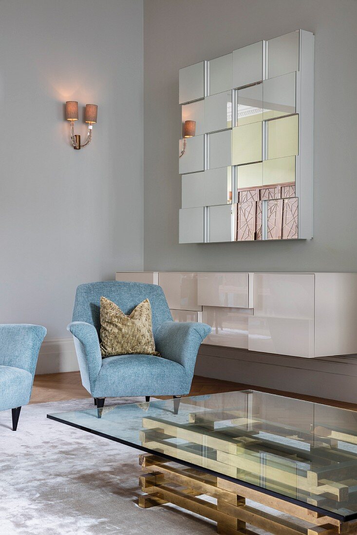 Glass coffee table with brass base, light blue armchairs and floating sideboard below mirrored wall-mounted cabinet