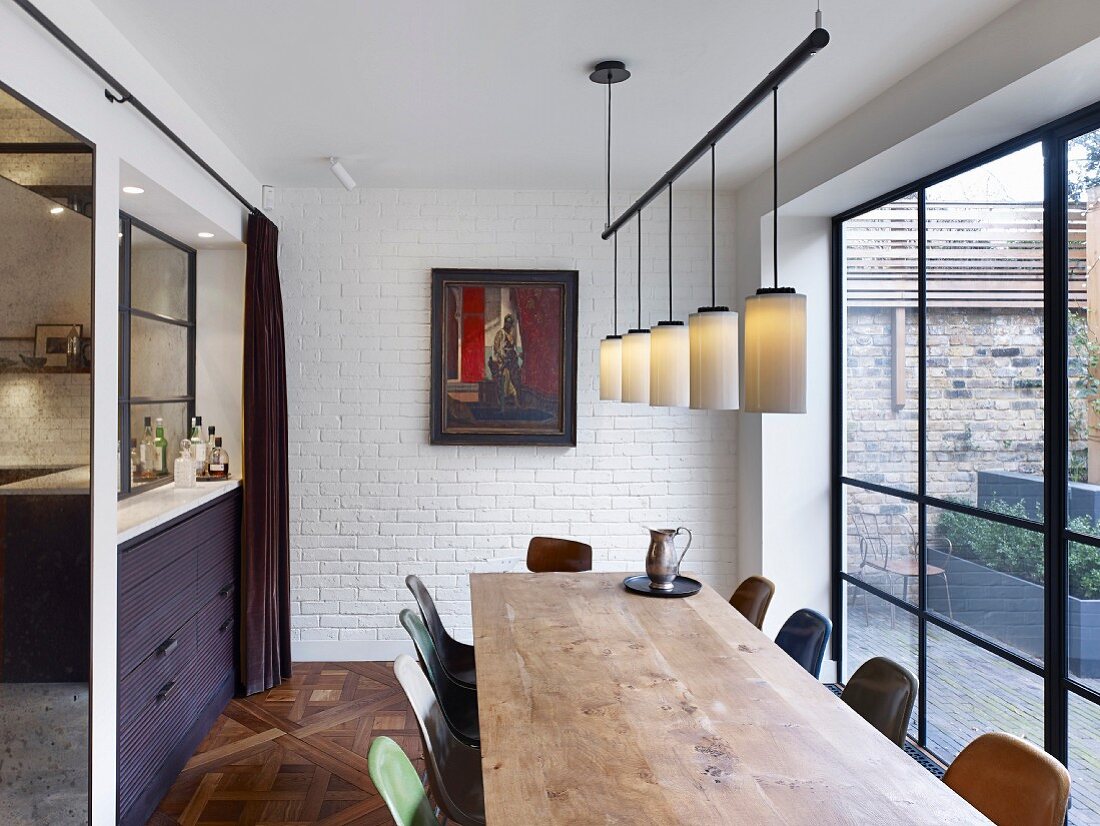 Long wooden table and retro chairs of various colours in dining room with view of terrace