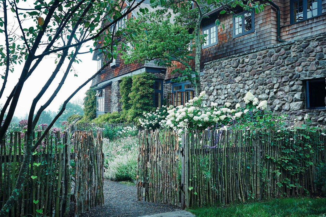 A stave fence with an open gate, white lilac bushes in front of an old country house with a natural stone and shingle facade