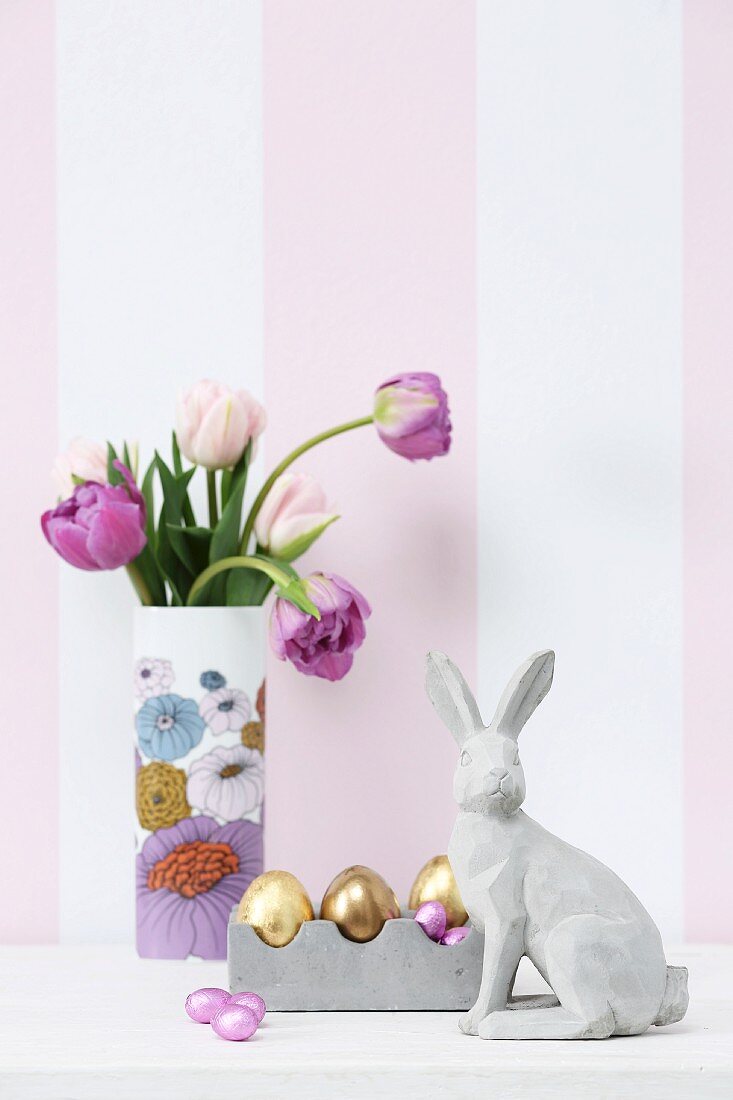 Concrete hare, Easter ornaments and vase of tulips against striped wallpaper