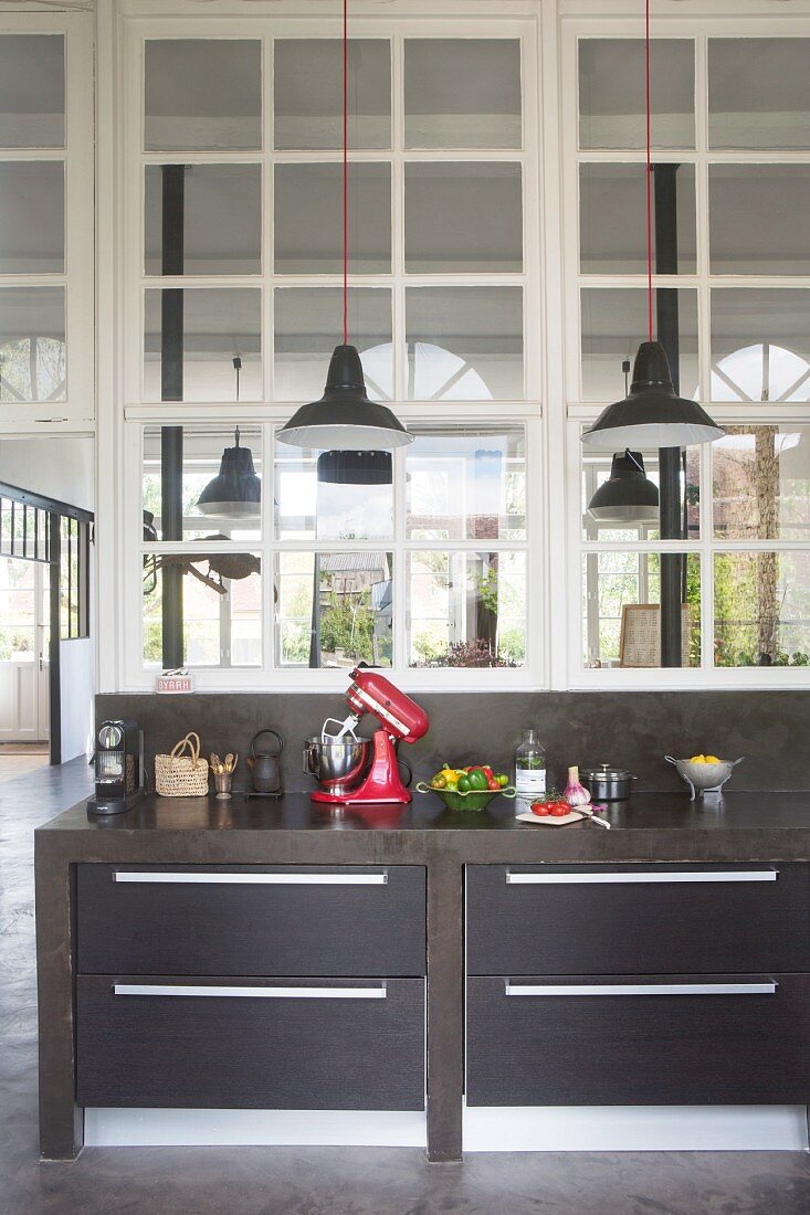 Black, concrete kitchen counter with drawers and kitchen utensils below interior windows in loft-style apartment