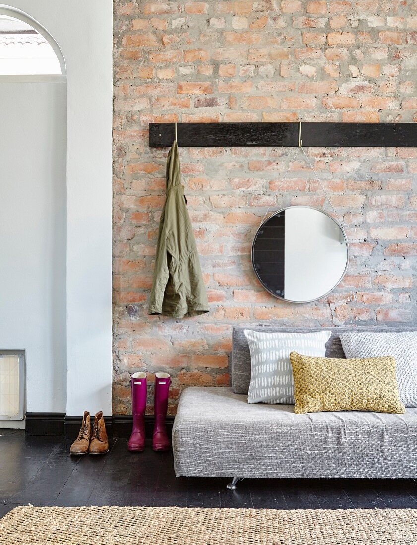 Grey sofa below coat rack and round mirror on brick wall