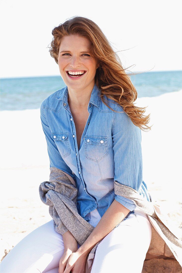 A young brunette woman on a beach wearing a denim shirt and a cardigan