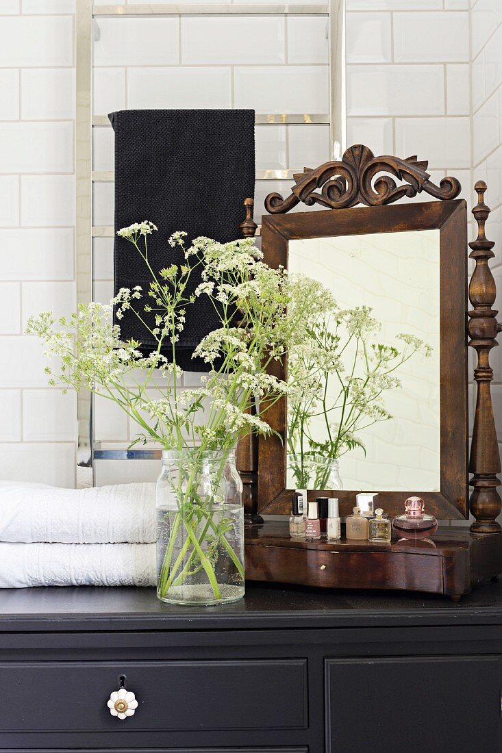 Antique vanity mirror, perfume bottles and glass vase of cow parsley on top of black bathroom cabinet