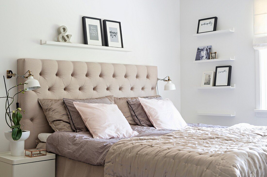 Double bed with beige, button-tufted headboard in white bedroom with pictures on narrow shelves