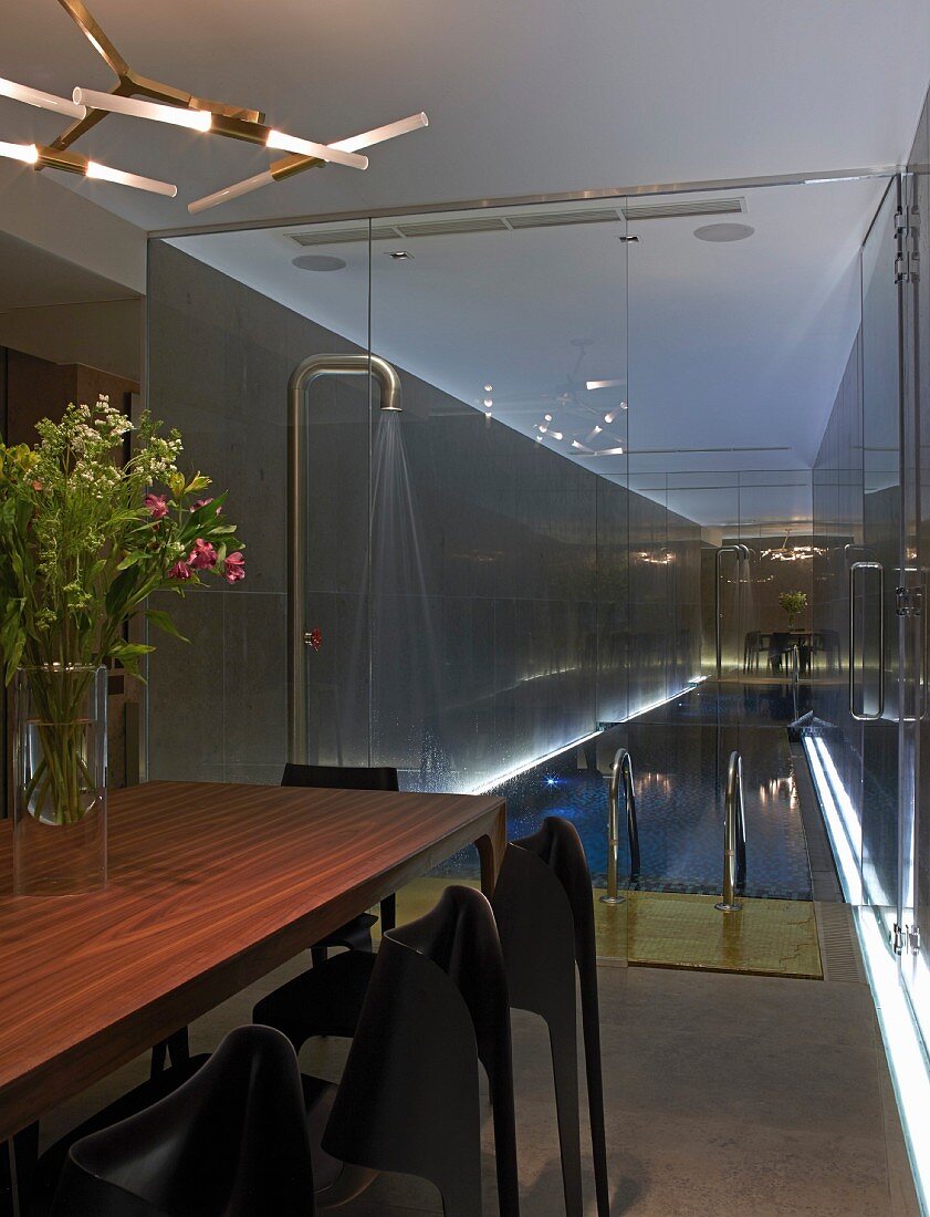 Exotic-wood table below designer lamp and next to glass partition wall with view of illuminated indoor pool and shower