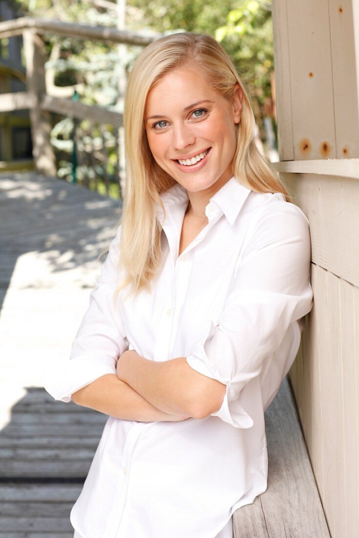 A young blonde woman leaning on a wooden facade wearing a white blouse