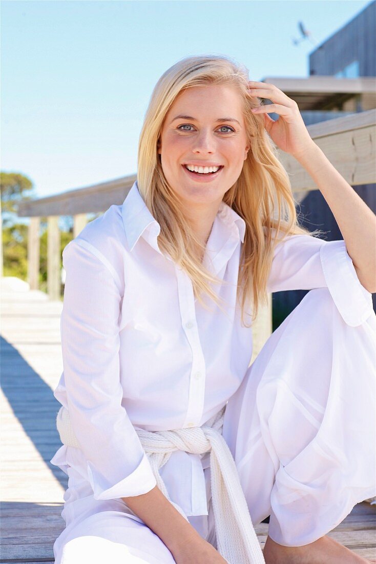 A young blonde woman sitting on a wooden jetty wearing a white blouse and trousers