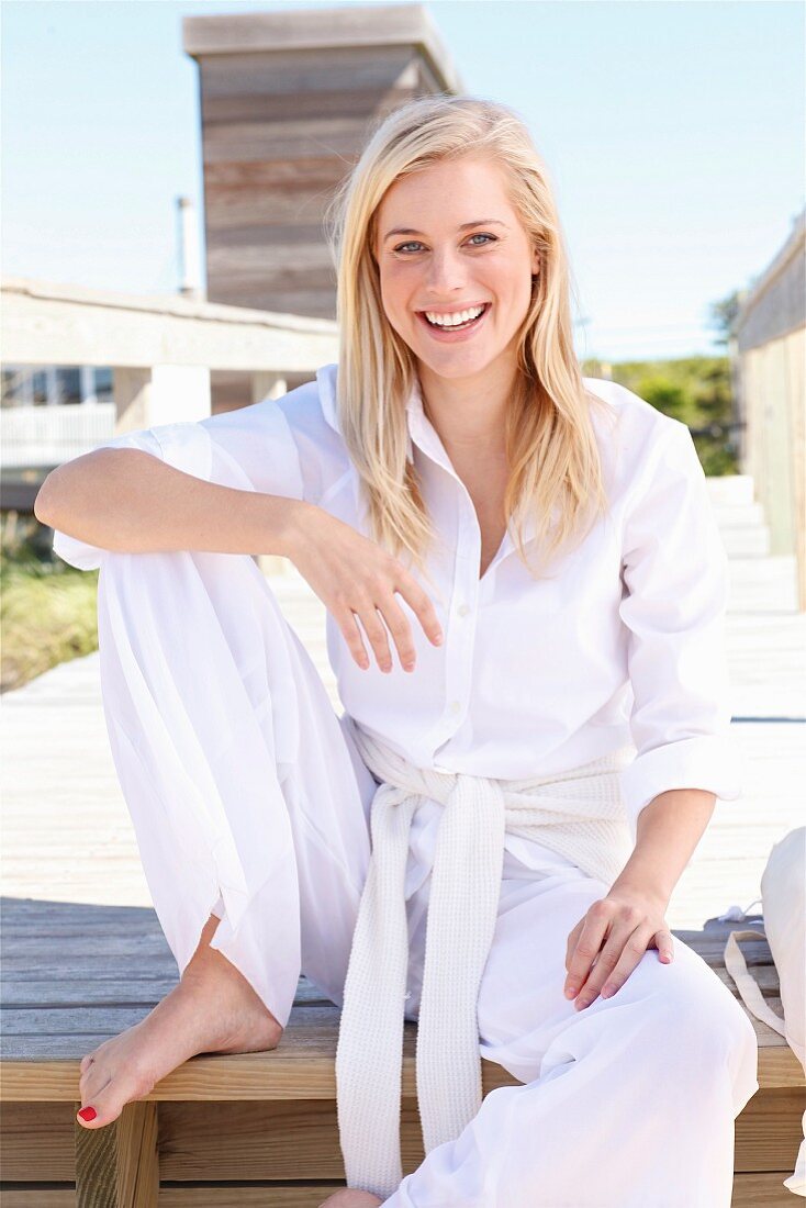 Junge blonde Frau in weisser Bluse und Hose sitzt an Holzsteg