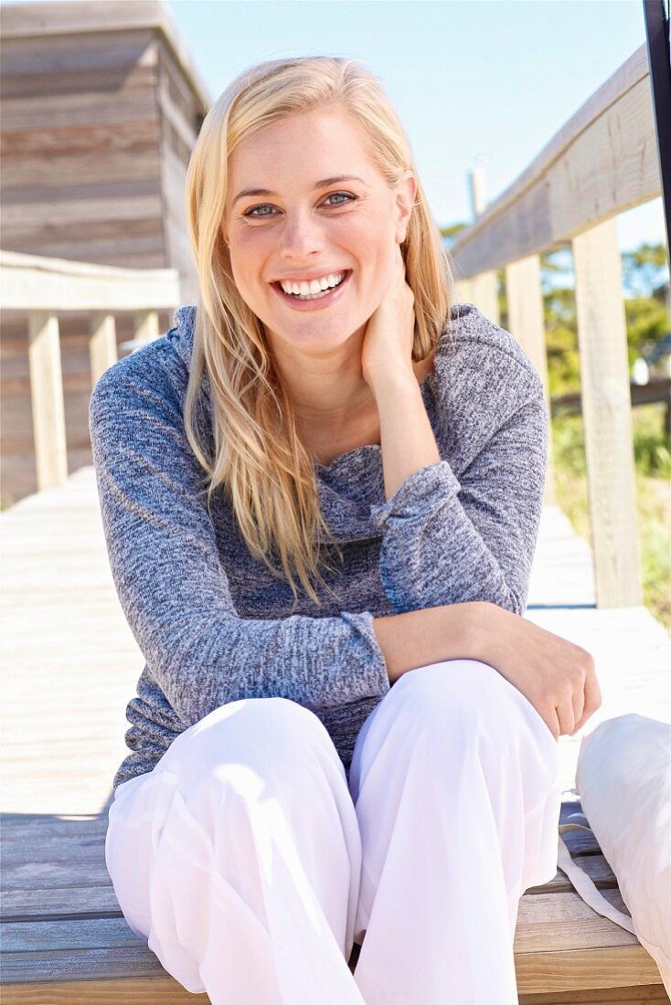 A young blonde woman on a wooden jetty wearing a grey jumper and white trousers