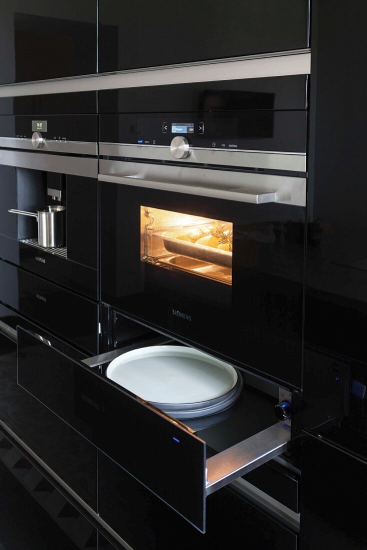 Küchenschrank mit schwarzer Hochglanzoberfläche, eingebauter Backofen und offene Wärmeschublade mit Tellern