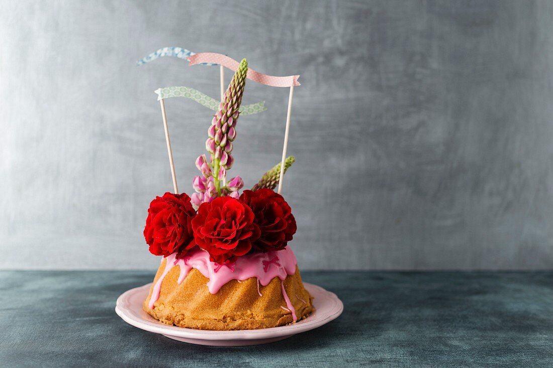 A mini Bundt cake decorated with icing, roses, Lupin flowers and paper flags