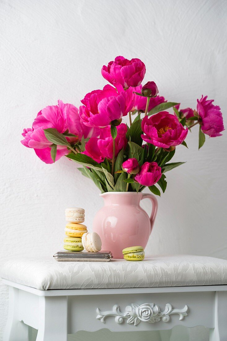 Macarons vor pinkfarbenem Pfingstrosenstrauss auf weißem Hocker