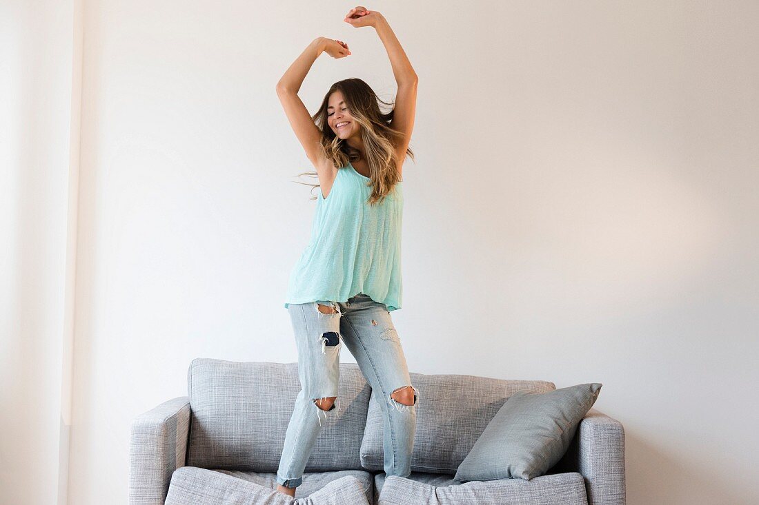 Junge Frau in zerrissener Jeans und türkisfarbenem Top tanzt auf Sofa