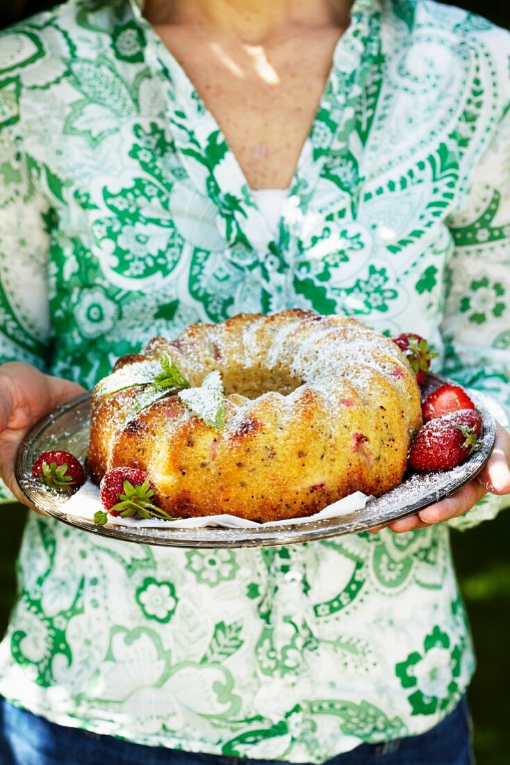 A woman holding a rhubarb and strawberry cake dusted with icing sugar