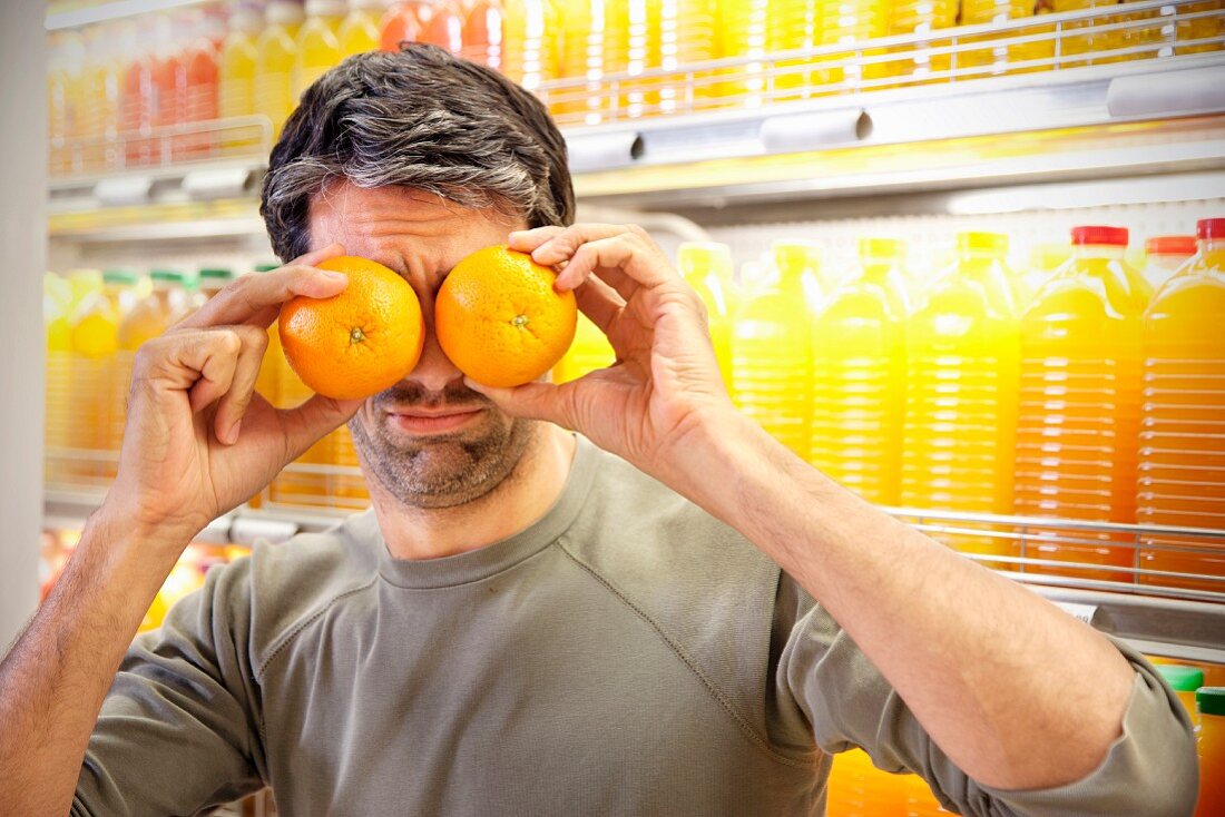 A man standing in front of a chiller cabinet with rows of juice bottles in a supermarket holding two oranges in front of his eyes