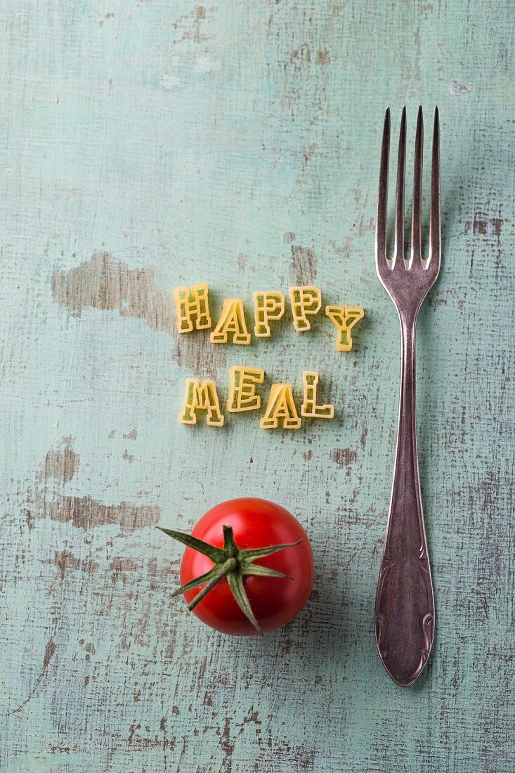 Schriftzug Happy Meal aus Buchstabennudeln daneben Gabel und Tomate