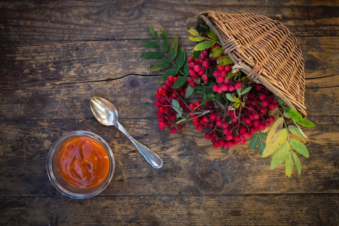 A jar of rowan berry jam next to a wicker basket of rowan berries (seen from above)