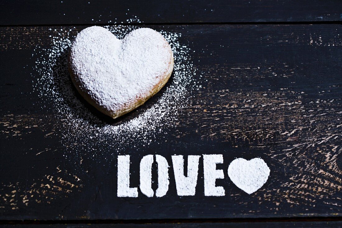 Herzförmiger Krapfen und das Wort Love aus Puderzucker auf Holzuntergrund