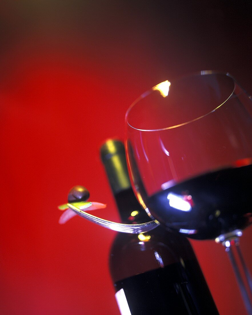 Red wine glass in front of a red wine bottle and fork with olive