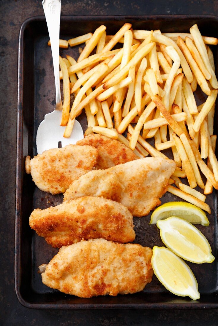 Chicken escalope, chips and lemon wedges on a baking tray