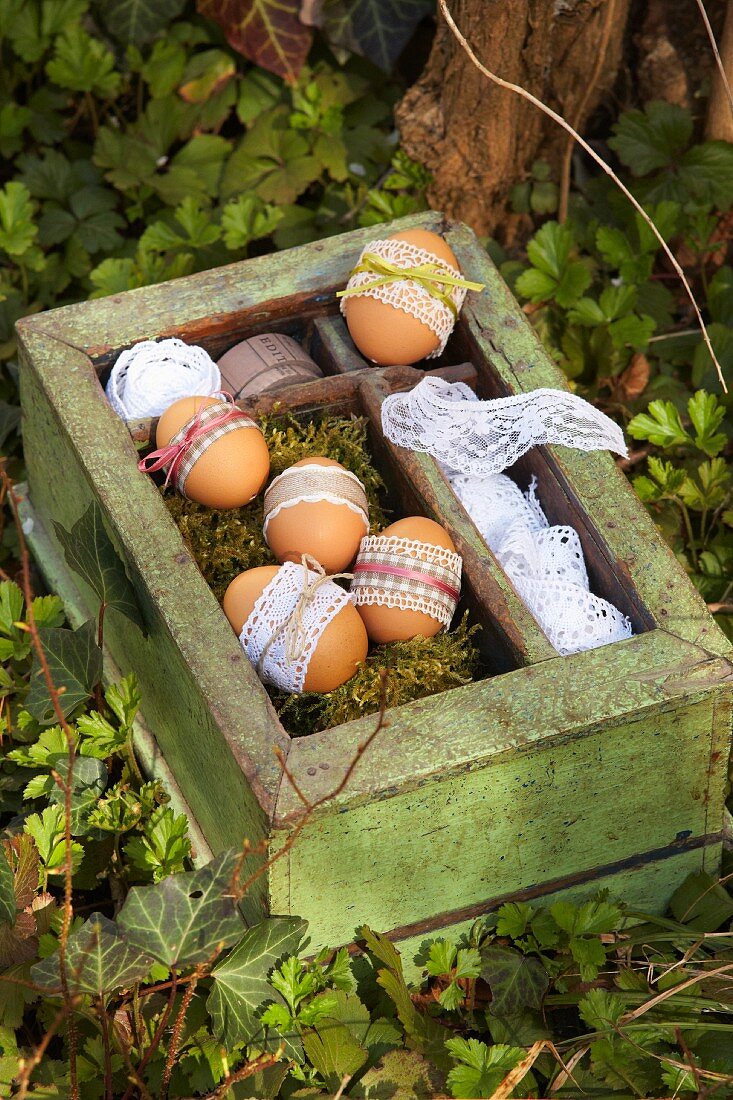 Ungefärbte Ostereier mit Spitzenbesatz und zartenSatinschleifchen dekoriert, in Vintage Holzkiste mit Moos
