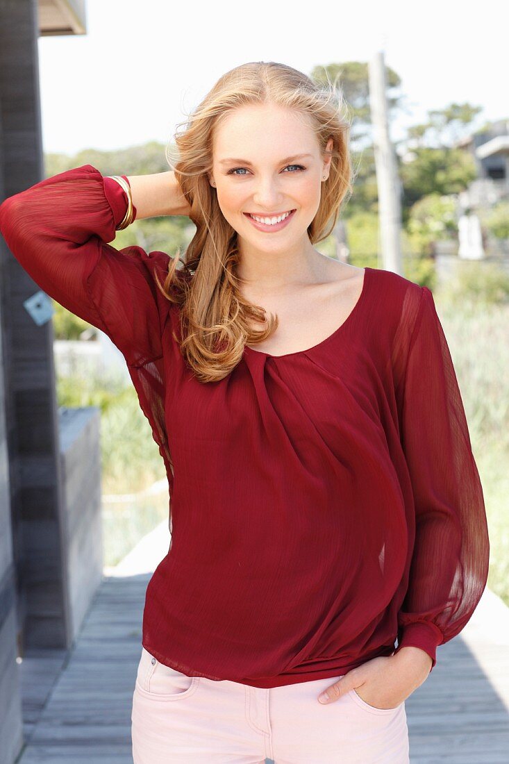 A young, dark blonde woman wearing a bordeaux red blouse and light trousers