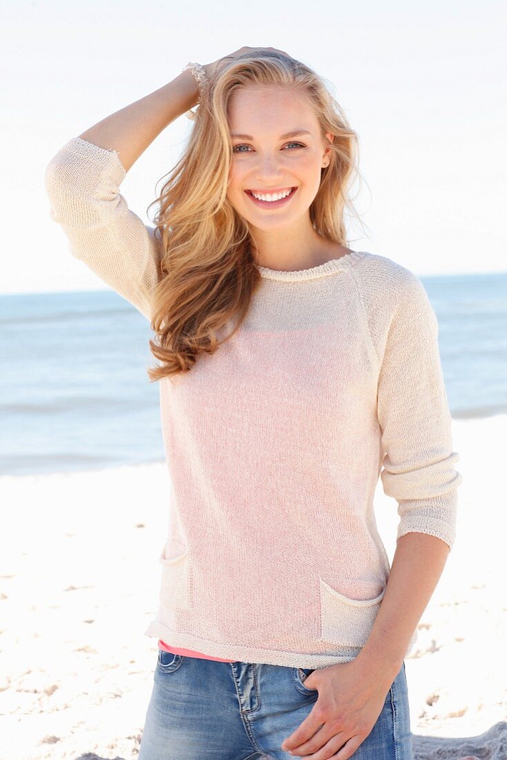 A young dark blonde woman on a beach wearing a light knitted jumper and jeans
